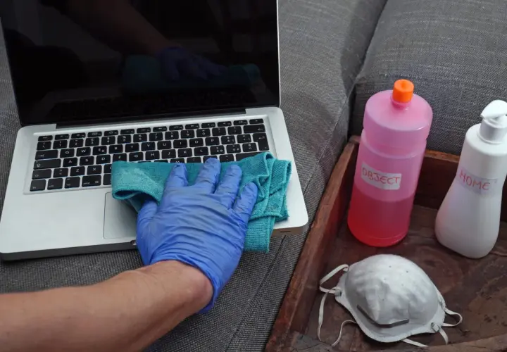 Ręka w niebieskiej rękawiczce czyści laptopa. Obok butelka z napisem "OBJECT" i maseczka. Denaturat jest do dezynfekcji przedmiotów.
