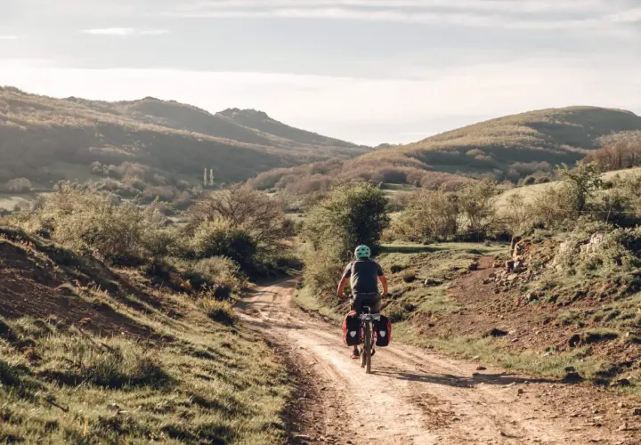 Ciclista pedaleando por un camino rural en España, disfrutando de sus viajes en bicicleta por España. Paisaje montañoso y vegetación verde.