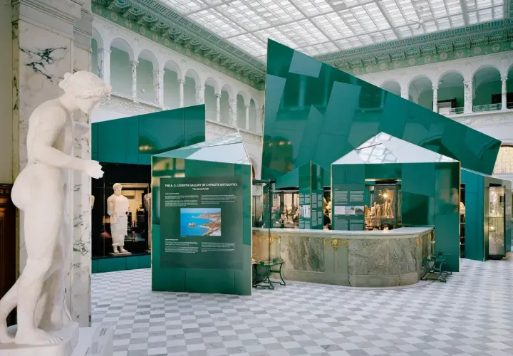 En marmorstaty i förgrunden på Medelhavsmuseet Stockholm. Gröna, geometriska installationer och antika föremål visas i den ljusa hallen.