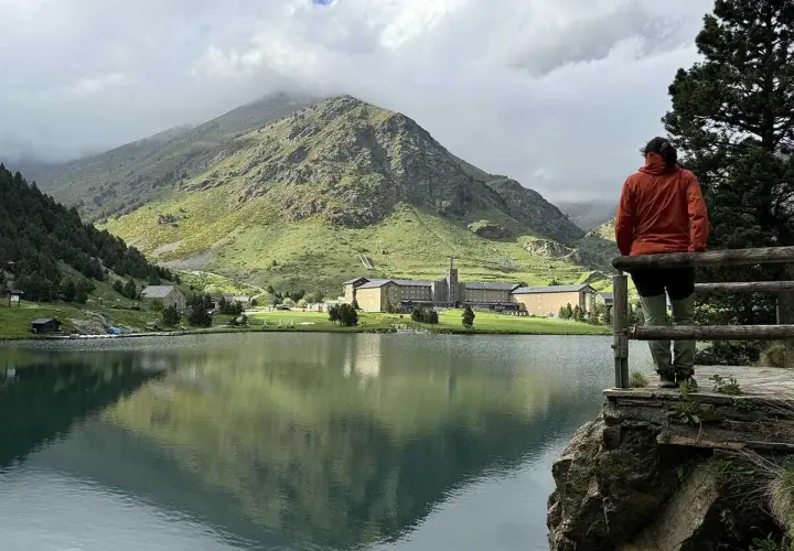 Persona contempla el sereno lago en el valle de Nuria, con imponentes montañas verdes y un complejo de edificios al fondo.