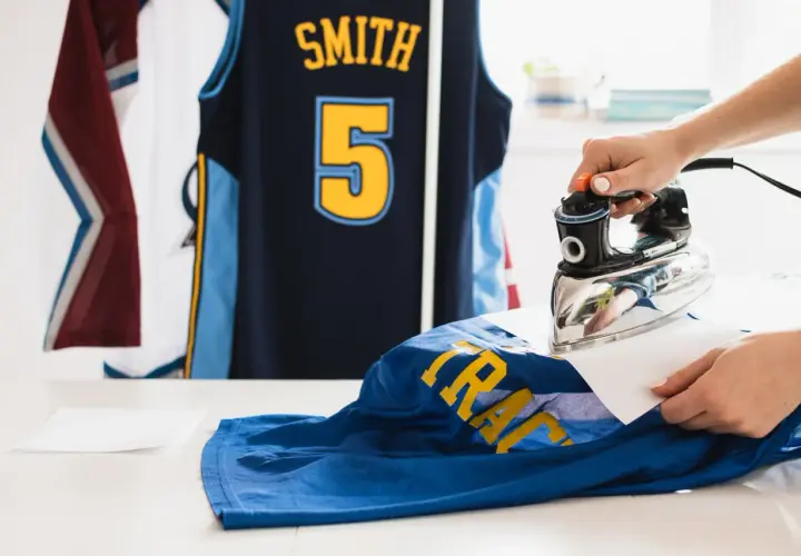 Une main utilise un fer à repasser pour défroisser un maillot de basket bleu avec des lettres jaunes. D'autres maillots sont suspendus en arrière-plan.