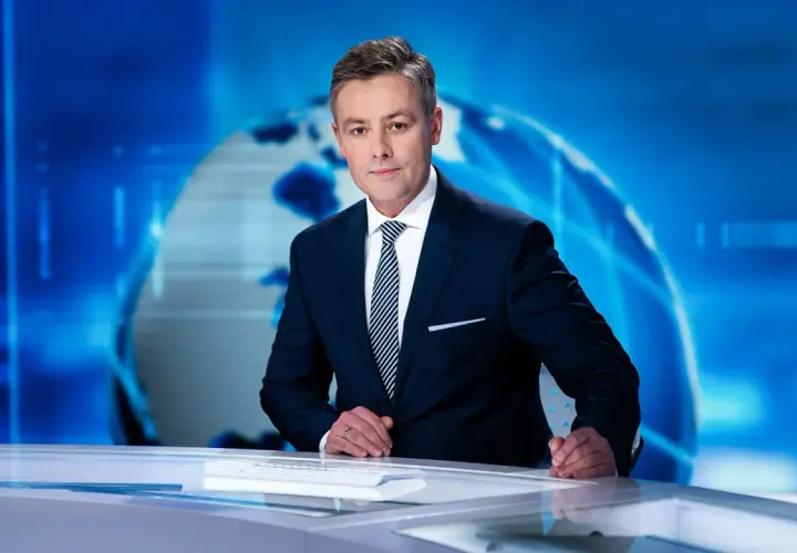 Elegancki konferansjer w garniturze stoi przy biurku na tle niebieskiej grafiki z globusem.