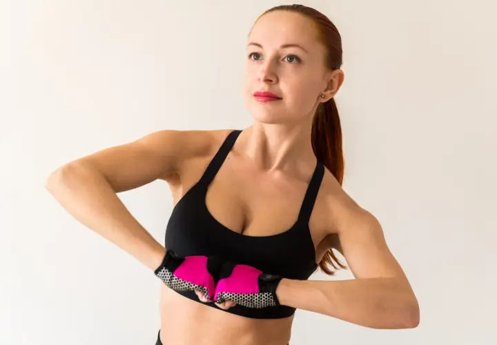 Kobieta w sportowym topie i rękawiczkach wykonuje ćwiczenia na podniesienie biustu, napinając mięśnie klatki piersiowej.