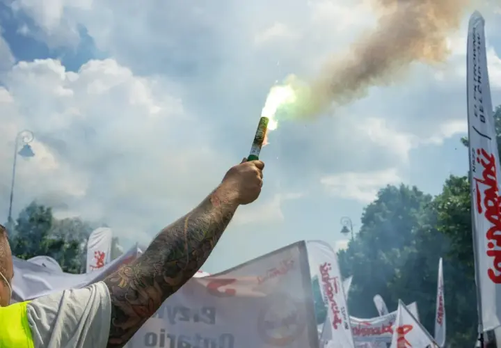 Dym z flary rozświetla niebo nad demonstracją związków zawodowych. W tle białe flagi z napisem "Solidarność".
