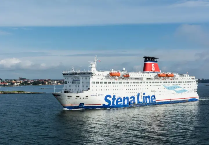 Statek Stena Line płynie po morzu, gotowy na rejs 24h. W tle widać wybrzeże z zabudowaniami.