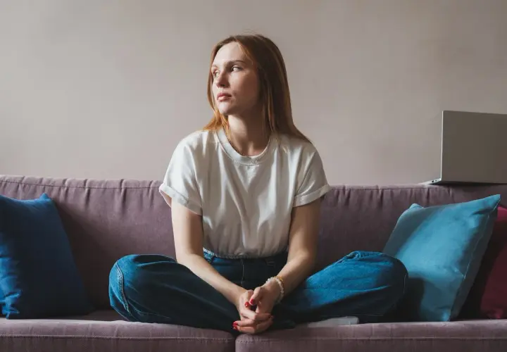 A young woman with red hair sits cross-legged on a couch, looking away thoughtfully.