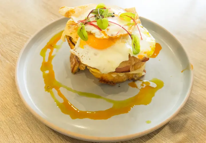Un délicieux croque madame avec un œuf au plat coulant, du jambon et du fromage fondu, garni de jeunes pousses et servi sur une assiette avec un filet de sauce.
