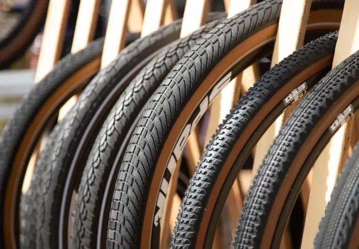 A row of diverse bicycle tires, featuring knobby mountain bike tires and smoother road bike tires, all with distinctive tan sidewalls, are displayed.