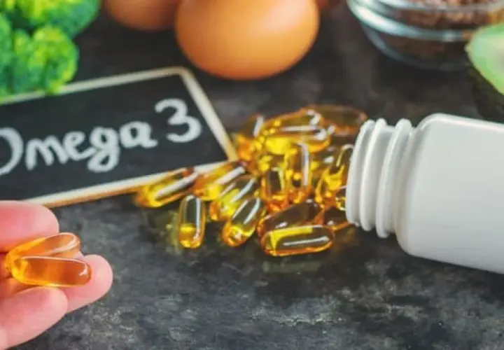 Kapsułki Omega-3 wysypujące się z białej butelki, na tle tabliczki z napisem "Omega 3".