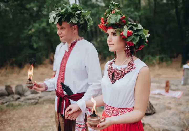 Para młoda w tradycyjnych strojach, z wiankami na głowach, trzymająca płonące świece. To piękna swadźba.