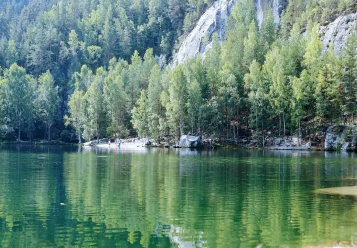 Zielone jezioro otoczone lasem i skalnymi formacjami. W oddali widać ludzi spacerujących po plaży. To malownicze miejsce, gdzie góry stołowe spotykają się z wodą.