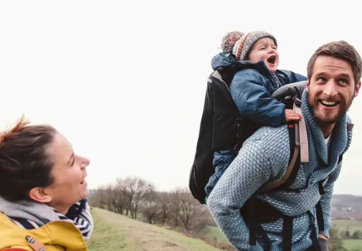 Szczęśliwa rodzina na spacerze. Tata niesie dziecko w nosidełku turystycznym, mama uśmiecha się do nich.