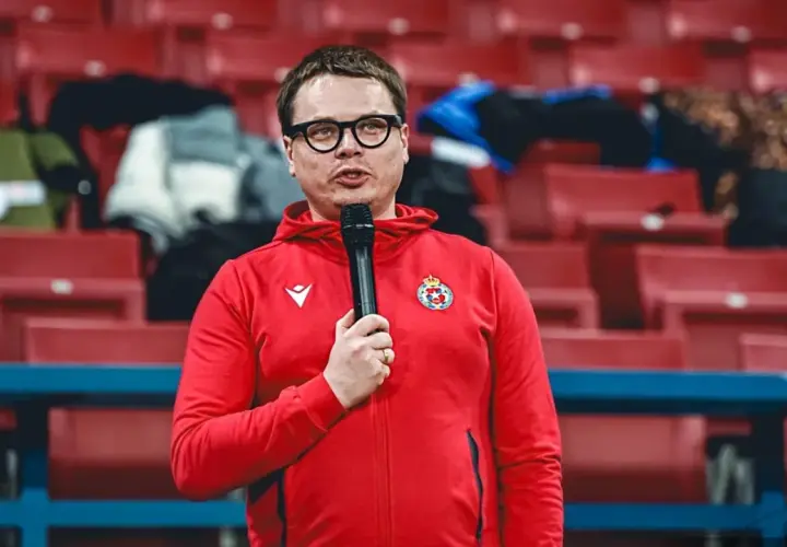 Właściciel Wisły Kraków w czerwonej bluzie z kapturem i mikrofonem, na tle pustych czerwonych trybun stadionu.