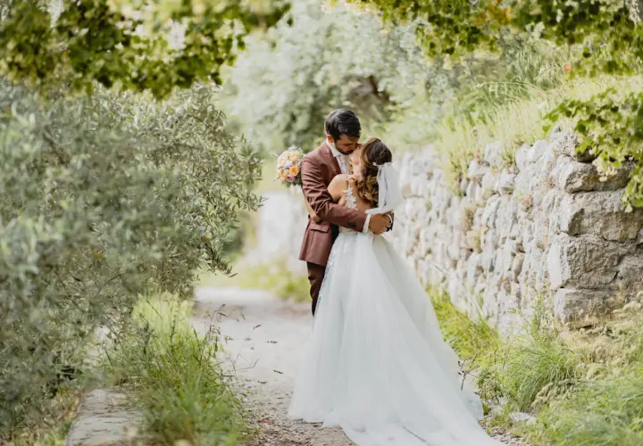 Un couple s'embrasse tendrement lors de leur mariage de 50 personnes, entouré par la nature verdoyante et un mur de pierres anciennes.