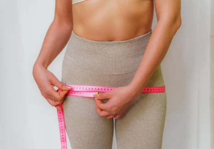 Mujer midiendo su cintura, observando la cinta métrica rosa que rodea sus caderas, enfocándose en los contornos y posibles hip dips.