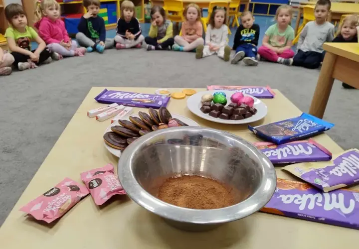 Dzieci w przedszkolu świętują dzień czekolady. Na stole czekolady Milka, Kinder, ciastka i miska z kakao.