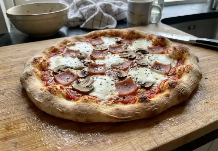 En gyllene, hemmagjord pizza med skinka och champinjoner ligger på en träskärbräda.