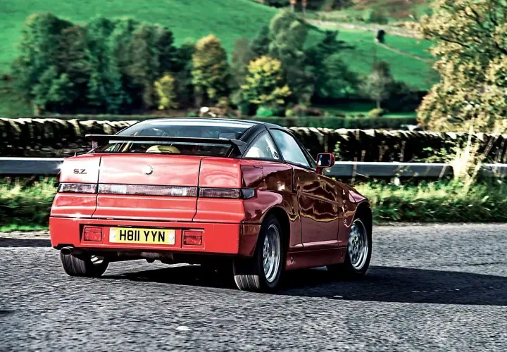 Czerwone Alfa Romeo SZ z czarnym dachem i spoilerem jedzie po wiejskiej drodze.