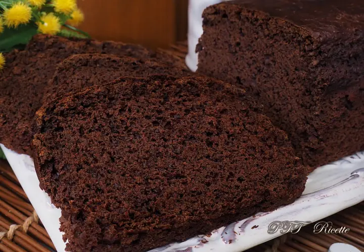 Plumcake al cacao sofficissimo, affettato su un piatto bianco, con fiori gialli sullo sfondo.