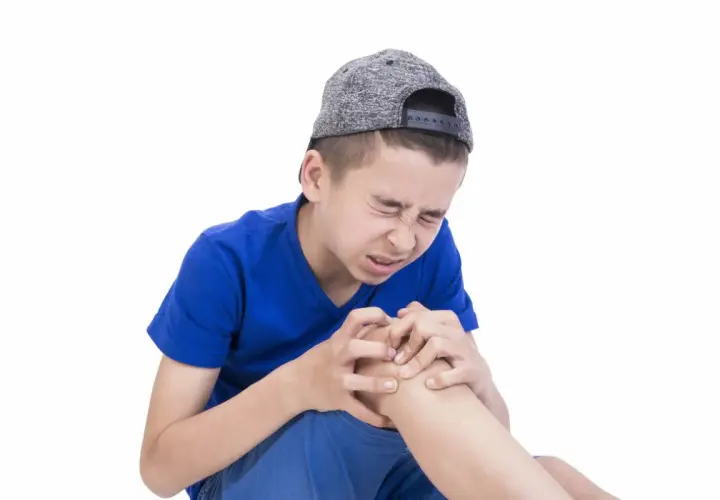 Ragazzo con cappellino grigio stringe il ginocchio con espressione sofferente, forse a causa dei dolori della crescita.