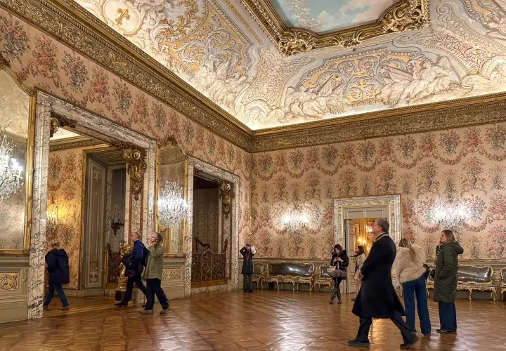 Elegancki przedpokój w pałacu z bogato zdobionym sufitem i tapetą w kwiaty. Zwiedzający podziwiają wnętrze.