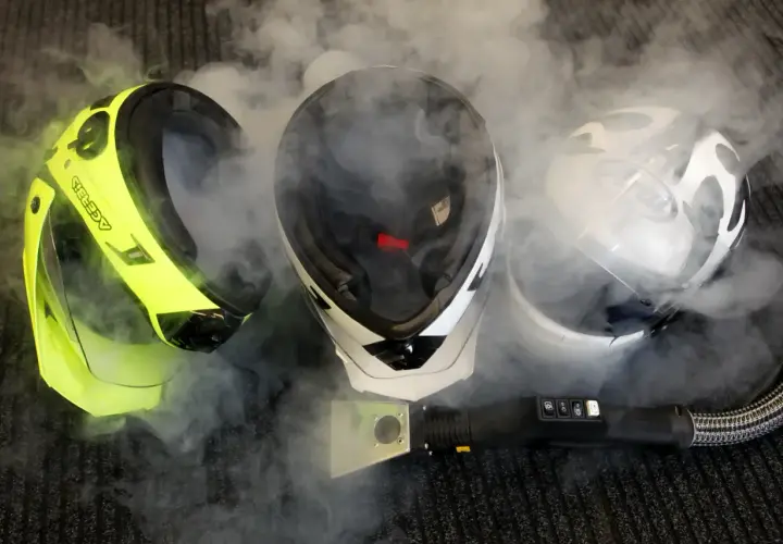Pulire interno casco moto con vapore. Tre caschi, uno giallo fluo, uno bianco e nero, uno bianco, circondati da fumo.