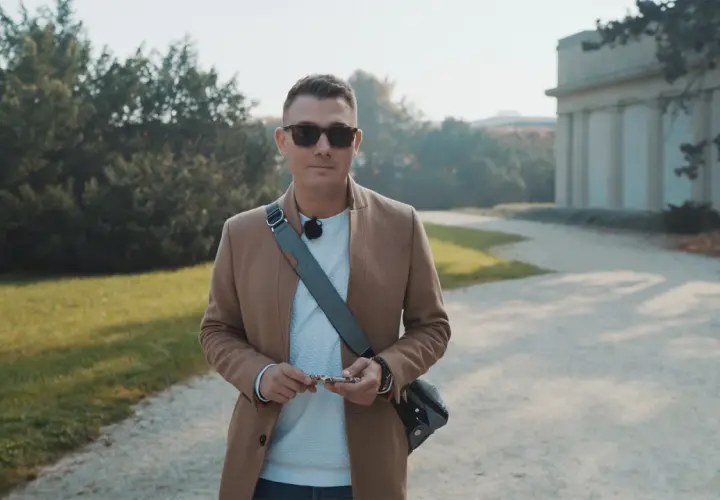 Mężczyzna w beżowym płaszczu i okularach przeciwsłonecznych, trzymający klucze, przygotowuje się do kursu fotografii telefonem w parku.