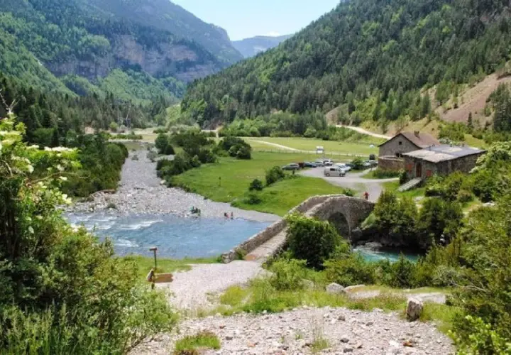 Un río de aguas turquesas fluye por un valle verde, cruzado por un puente de piedra. La ruta Bujaruelo invita a disfrutar de este paisaje montañoso.