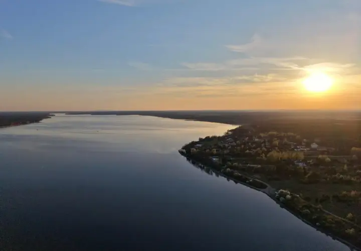 Zachód słońca nad Zalewem Sulejowskim. Spokojna tafla wody odbija niebo, a na brzegu widać domki letniskowe i las.