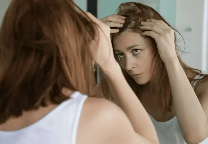 Mujer preocupada revisa su cuero cabelludo en el espejo, buscando soluciones sobre como frenar la caida del cabello.