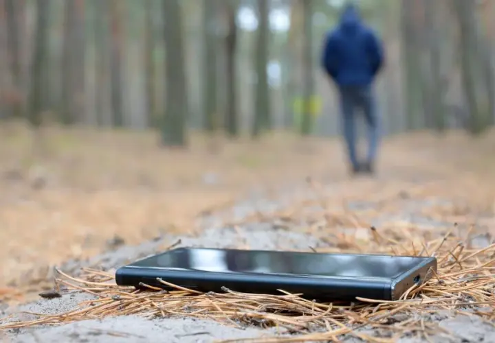 Ein Handy verloren, was tun? Ein schwarzes Smartphone liegt im Wald auf Nadeln. Im Hintergrund geht eine Person weg.