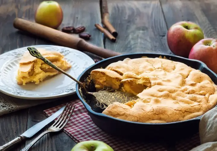 Una deliziosa torta di mele in padella, appena sfornata, con una fetta servita su un piatto. Mele fresche e cannella completano la scena rustica.