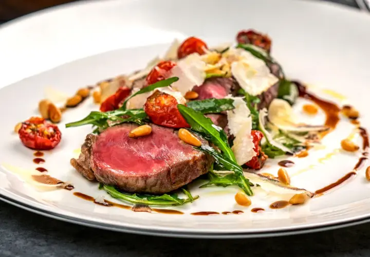 Saftiges Tagliata di Manzo mit Rucola, Kirschtomaten, Pinienkernen und Parmesan auf einem weißen Teller.