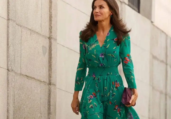La reina Letizia, con un vestido verde estampado y pendientes llamativos, camina con elegancia junto a un muro de piedra.