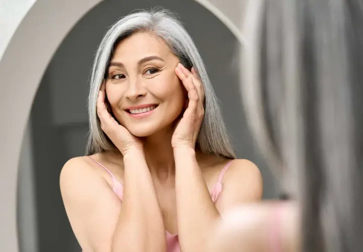 Une femme souriante aux cheveux gris, se touchant le visage, cherche à combler les rides naturellement.