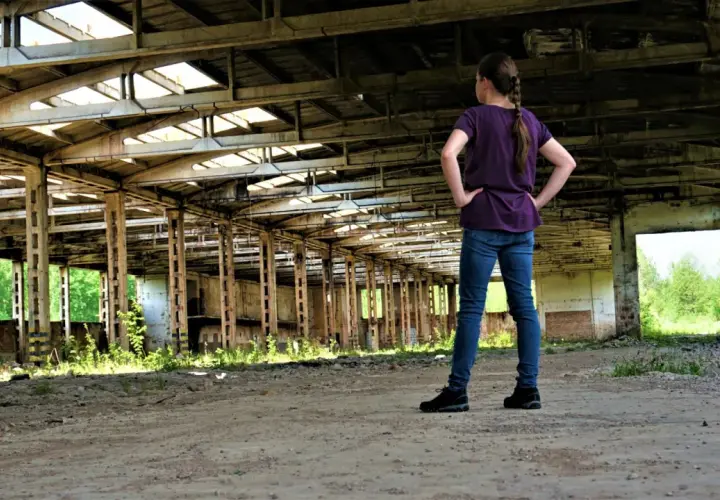 Dziewczynka w jeansach i fioletowej bluzce stoi w opuszczonej hali, odkrywając tajemnice urbex.