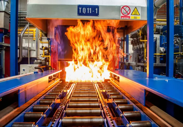 Płomienie buchają z pieca, w którym odbywa się nawęglanie stali. Taśmociąg z rolkami transportuje metal przez gorący proces.