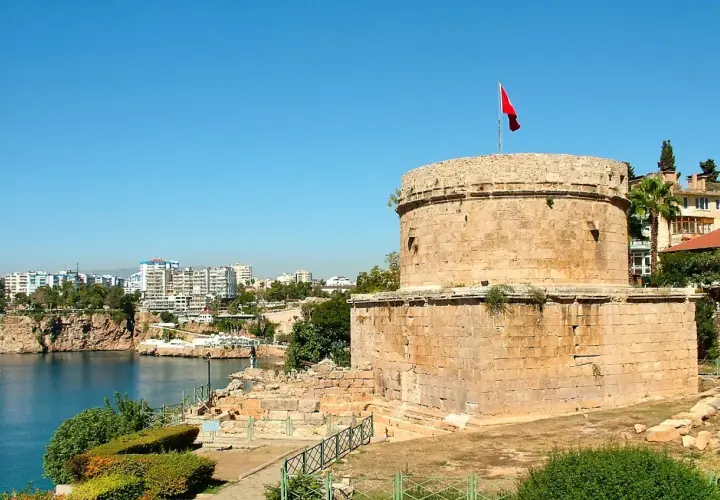Antalya Altstadt: Ein runder Steinturm mit türkischer Flagge überragt die Bucht. Im Hintergrund moderne Gebäude und Felsen.