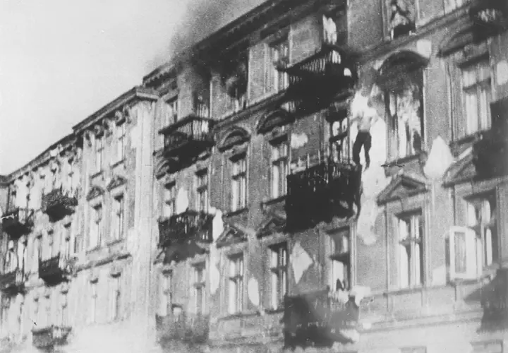 Brennende Gebäude im Warschauer Ghetto. Rauch steigt auf, Fenster sind zerbrochen, Balkone sind dunkel.