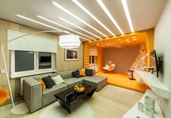 Stort vardagsrum med en grå soffa, svart soffbord och en orange sittgunga. Taket har en unik design med många ljuslister.