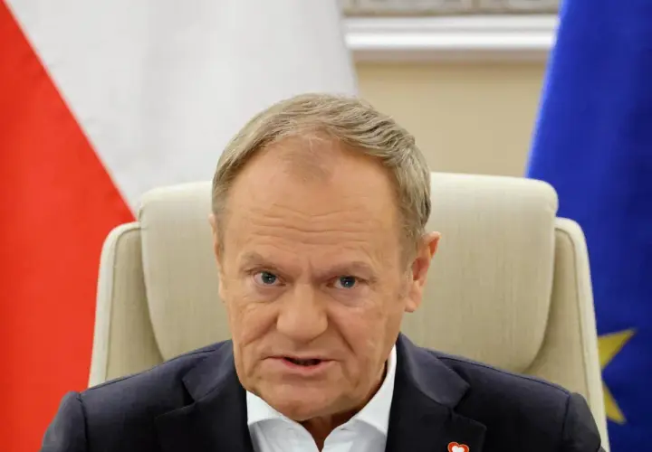 Donald Tusk, z flagą Polski i UE za plecami, w białej koszuli i granatowej marynarce. Czy Donald Tusk ma pochodzenie niemieckie?