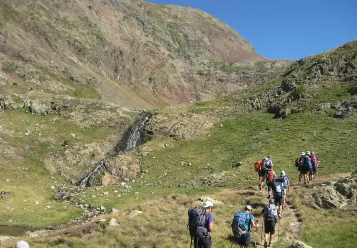 Senderistas caminan por un sendero de montaña hacia una cascada, como si fuera la porta del cel.