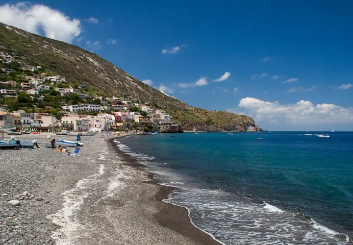 Vue d'une plage de galets sur l'île de Lipari, avec des maisons colorées accrochées à la colline et des bateaux dans la mer d'un bleu profond.