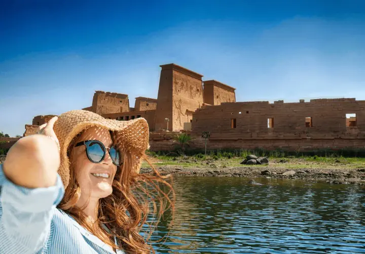 Mujer con sombrero y gafas de sol sonríe en un barco, con templos egipcios de fondo. ¡La mejor época para viajar a Egipto es ahora!