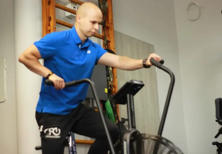 Mężczyzna w niebieskiej koszulce wykonuje ćwiczenia aerobowe na rowerze stacjonarnym, trenując wytrzymałość.