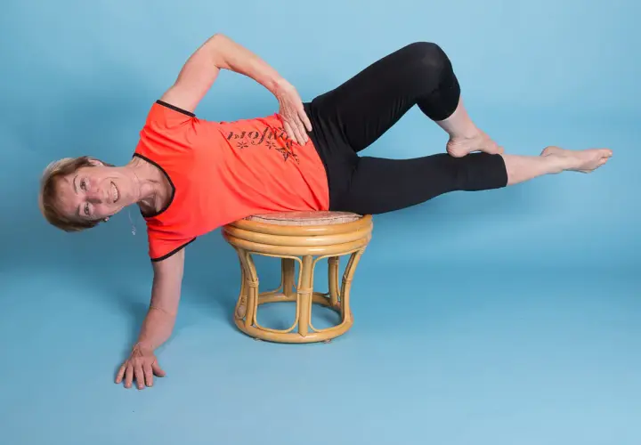 Ältere Frau in orangefarbenem Shirt und schwarzen Leggings macht eine seitliche Übung auf einem Hocker. Ihr linker Unterschenkel ist angewinkelt, der rechte gestreckt.
