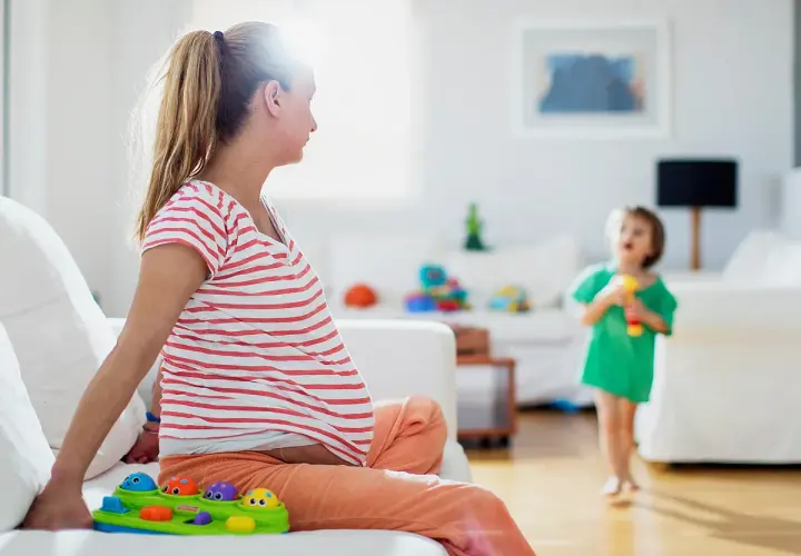 Schwangere Frau sitzt auf Sofa, Kind spielt. Frage: haushaltshilfe nach op wie viele stunden?