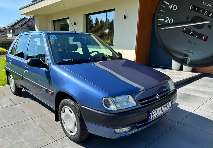 Niebieski Citroen Saxo z niskim przebiegiem, zaparkowany przed nowoczesnym domem. Na desce rozdzielczej widać licznik z przebiegiem 14649 km.