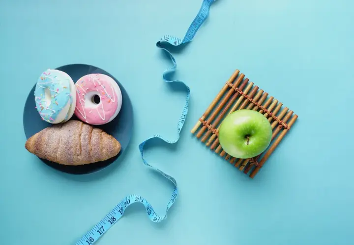 Dwa pączki i rogalik na talerzu obok zielonego jabłka i miarki krawieckiej. Wybór między słodyczami a zdrowiem, między kj a kcal.