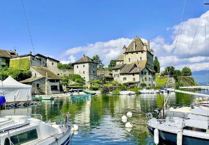 Vue d'une aire de camping-car au bord du lac Léman, avec un château médiéval, des bateaux et des maisons en pierre.